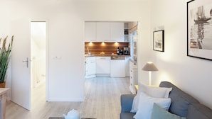 A modern living room with a grey sofa, a coffee table with cups and a teapot, a kitchen in the background with white cabinets and wooden countertops, and a framed picture on the wall.