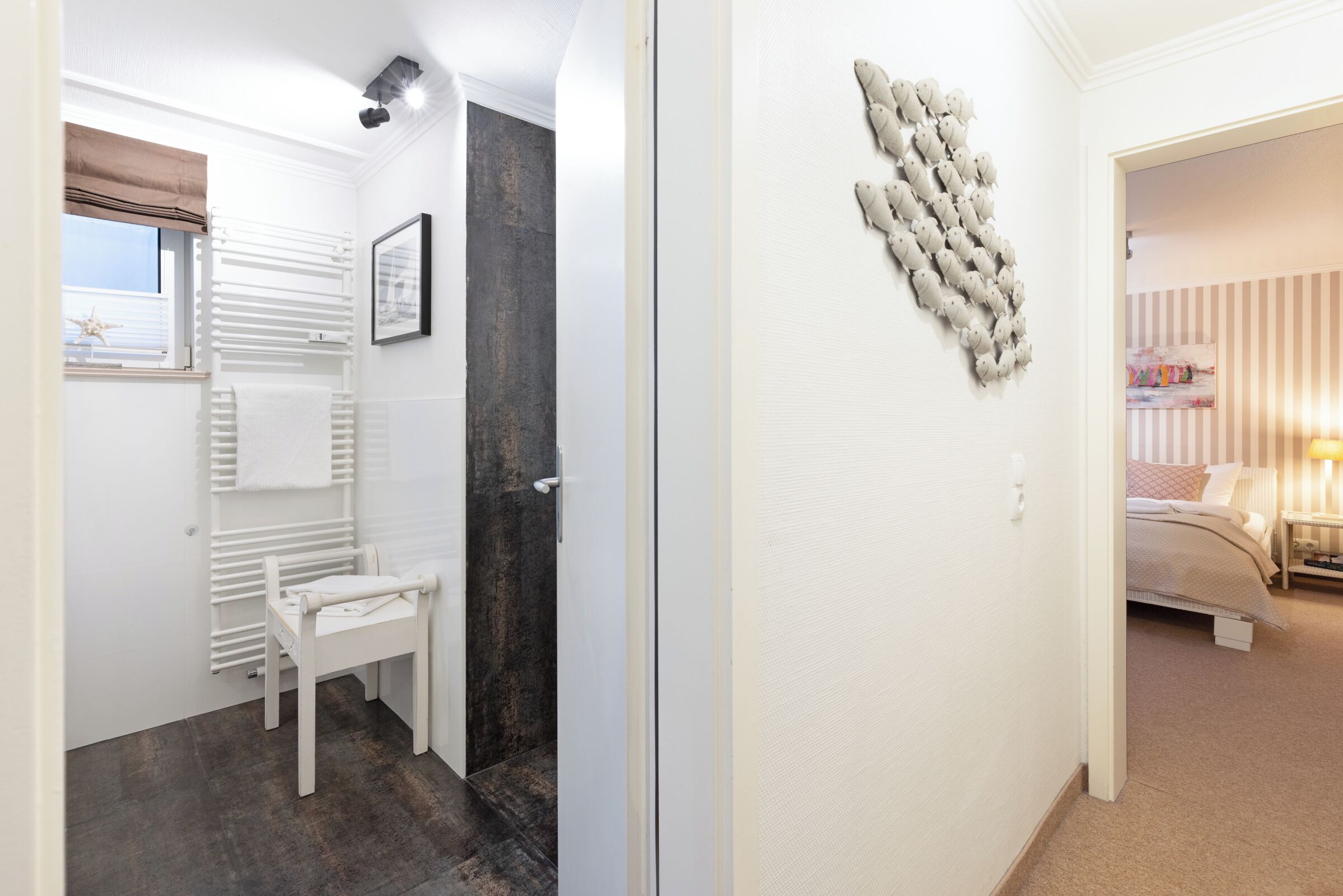 A hallway with a white radiator, a white chair, a dark wall, and a door leading to a bedroom with a bed and a nightstand.