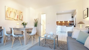 A modern living room with a grey sofa, a glass coffee table, and a dining area with a white table and chairs. A kitchen is visible in the background.