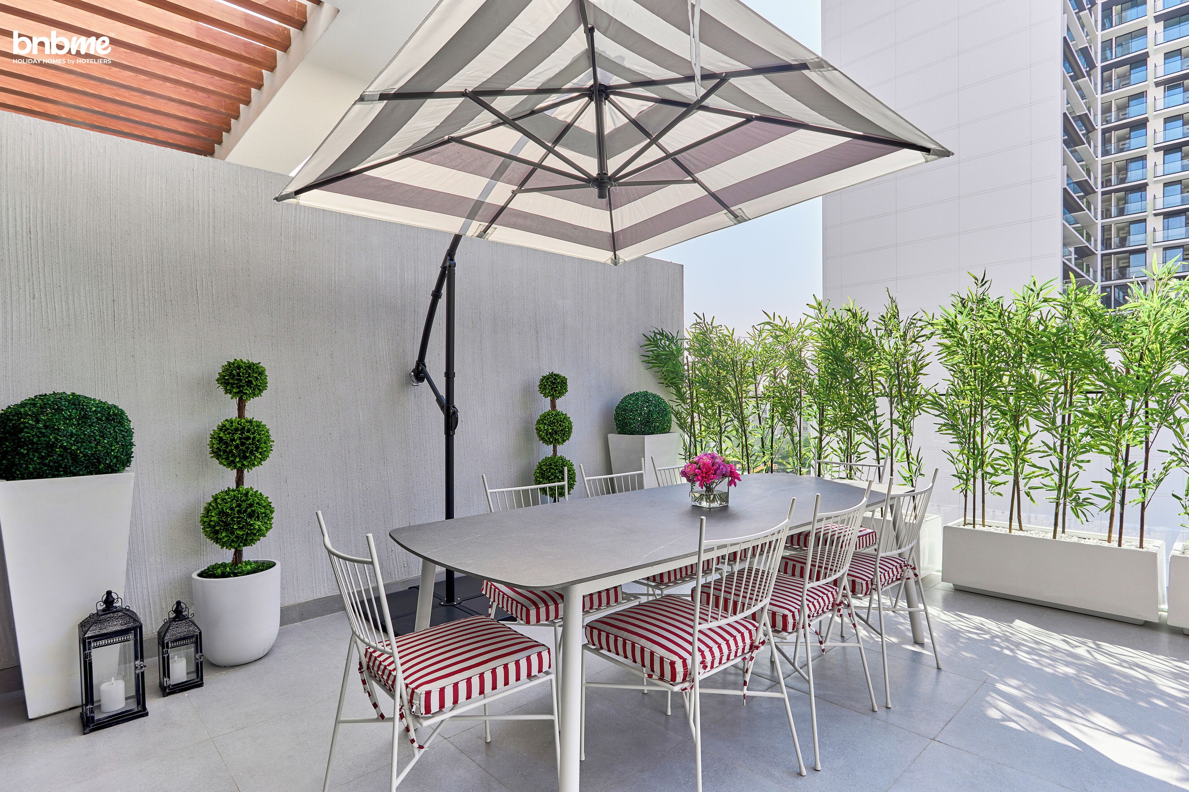 Een modern buitenontbijtgedeelte met een grote parasol, witte stoelen met kussens in het rood-wit gestreept patroon, en keurig opgestelde potplanten.