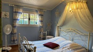 Un dormitorio con cama, ventilador de pie, ventana con cortinas y lámpara de pared.