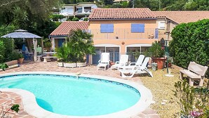 Una piscina con borde curvo, rodeada por un patio de piedra. Se observan sillones blancos y una pequeña mesa.