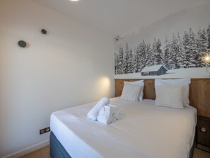 A neatly made bed with white linens and towels, a wooden headboard, and a wall-mounted painting of a snowy forest scene.