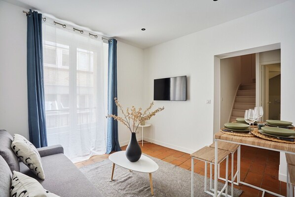 Un saló modern amb un sofà, una taula de centre amb un ram i una zona de menjador amb una taula preparada per a dues persones, i una escala que porta a un altre pis.
