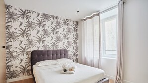 A bedroom with a bed, a headboard, a window with curtains, and a wall with a floral pattern. - Charming Duplex - Louvre Museum (Paris)
