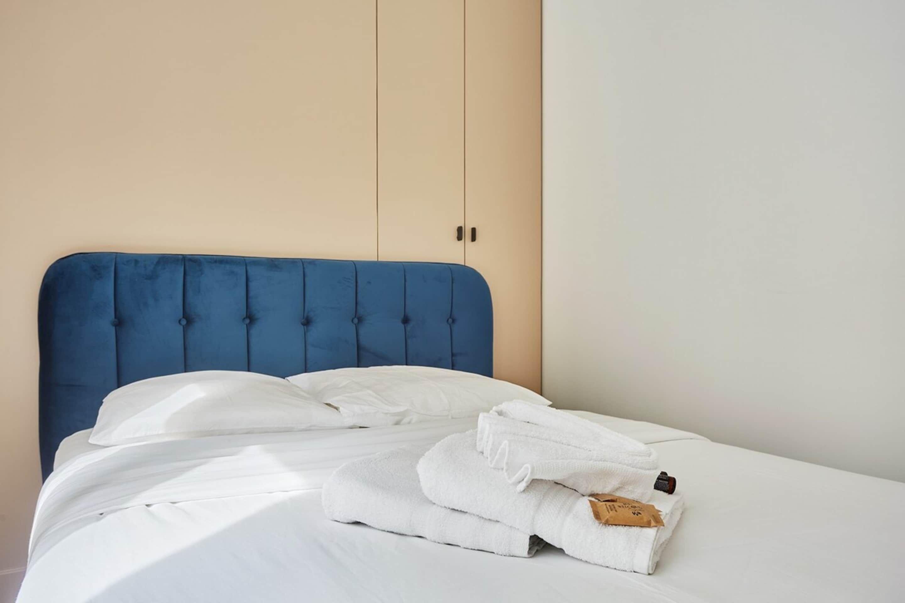 Een bed met een blauw hoofdeinde, witte beddengoed en een netjes gevouwen handdoek ernaast.