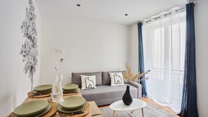 A modern living room with a grey sofa, two cat-themed cushions, a small round table, and a vase with dried plants. - Charming Duplex - Louvre Museum (Paris)