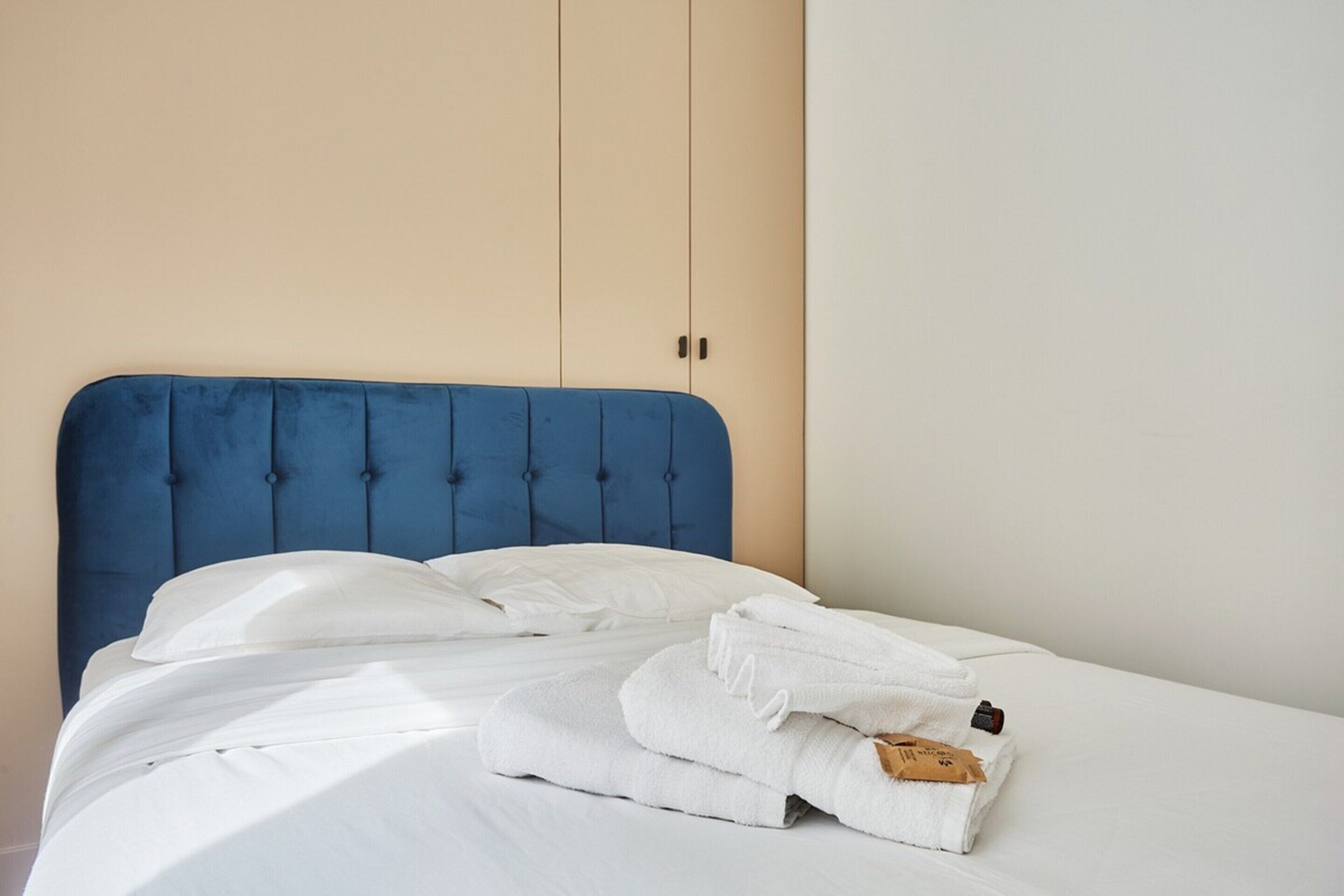 A neatly made bed with white linens and a blue headboard, accompanied by folded towels.