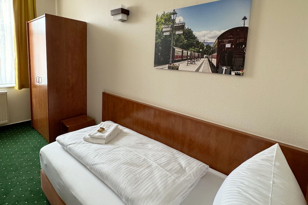 A hotel room with a bed, a wooden wardrobe, a wall-mounted lamp, and a picture of a train station.