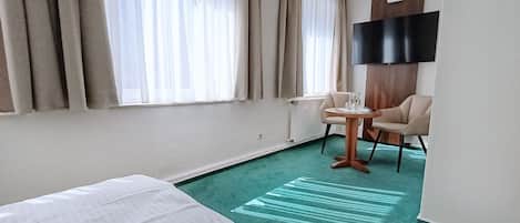 Habitación de hotel con cama, una pequeña mesa redonda con sillas, televisor de pantalla plana en la pared y ventana con cortinas.