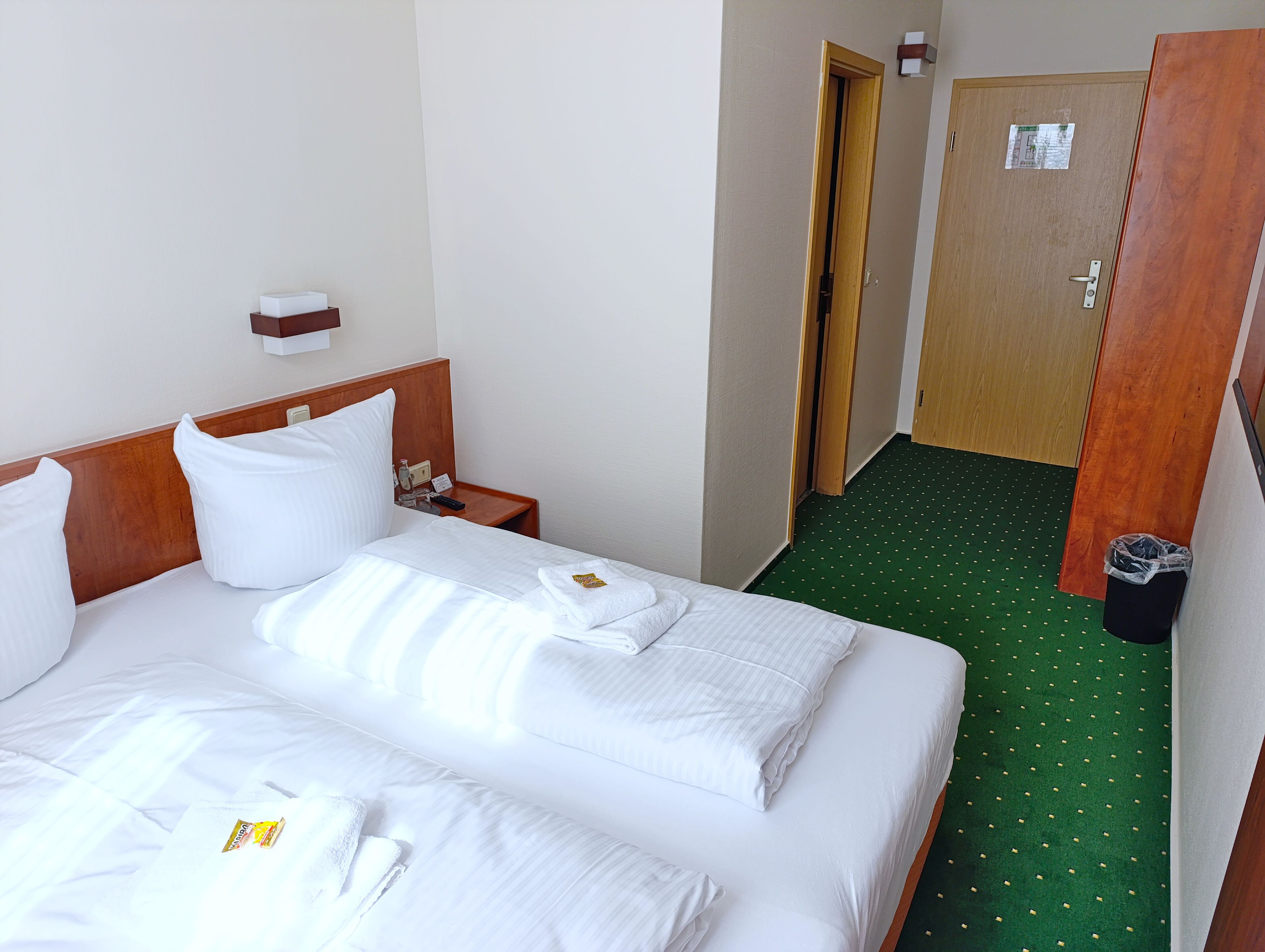 A hotel room with a neatly made bed, a wooden headboard, a nightstand with a lamp, a small table, and a green carpeted floor.