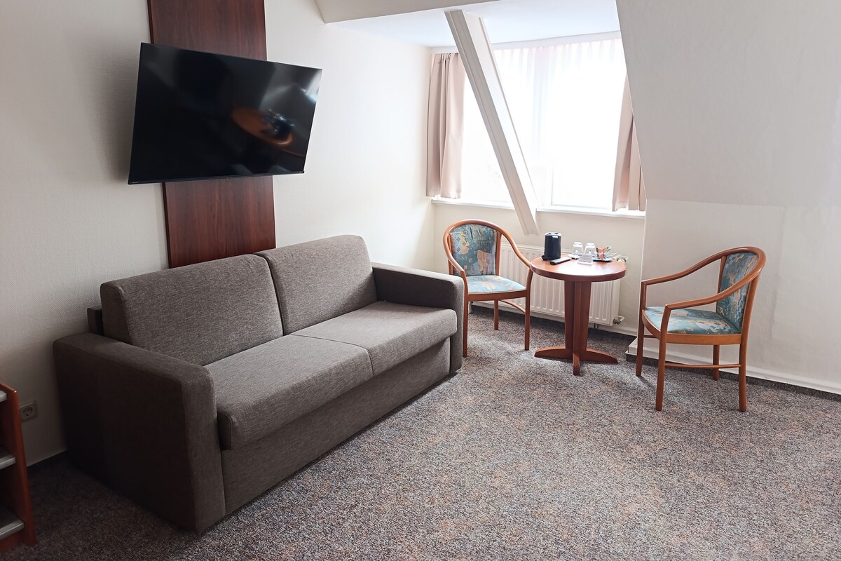 Een moderne hotelkamer met een grijze bank, twee houten stoelen, een klein rond tafeltje en een flatscreen televisie aan de muur.