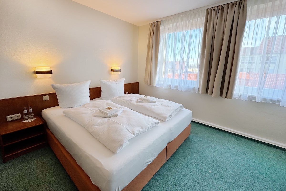 A hotel room with a large bed, two bedside tables, a nightstand, a window with sheer curtains, and a view of buildings outside.