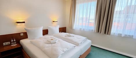 A hotel room with a large bed, two bedside tables, a nightstand, a window with sheer curtains, and a view of buildings outside.