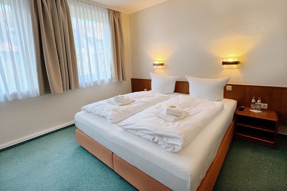 A hotel room with a large bed, two bedside tables, a nightstand, and a window with curtains.