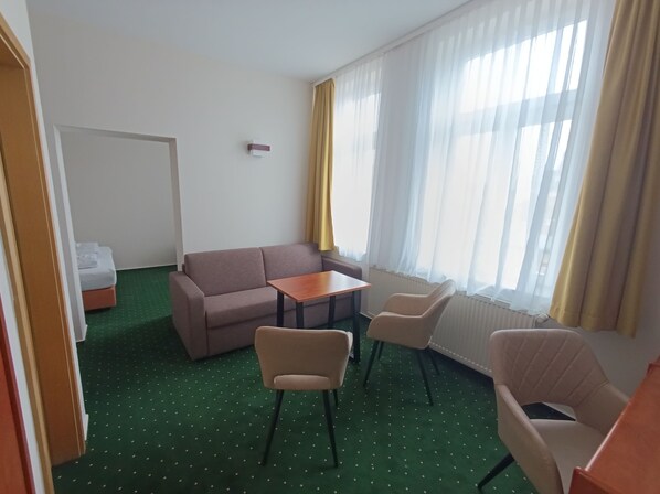 Ett hotellrum med ett grönt golv, en soffa, två fåtöljer, ett litet bord och ett fönster med gardiner.