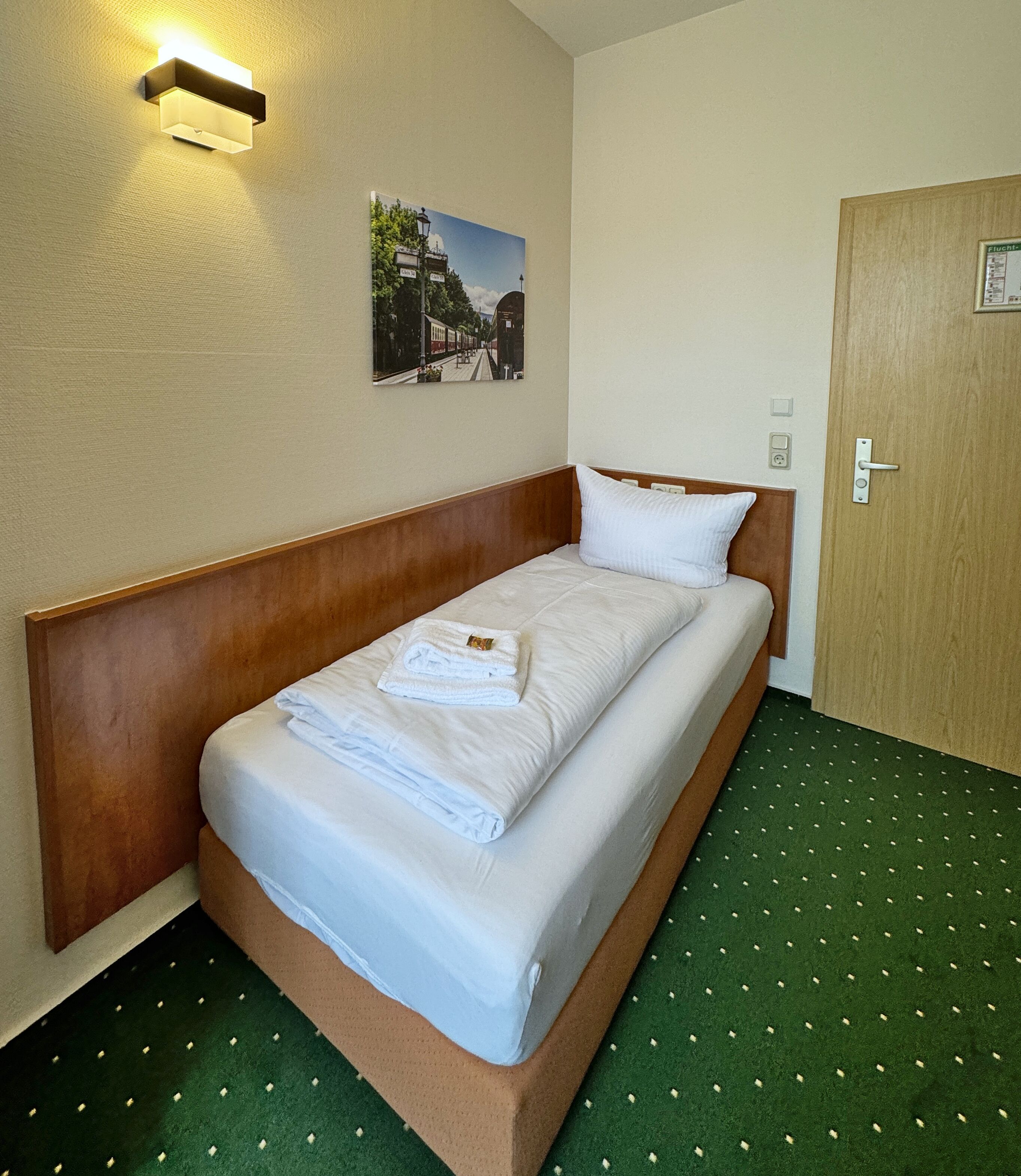A single bed with a wooden headboard, a neatly made bed with white linens and a folded towel, a green carpet with a dotted pattern, a wall-mounted light fixture, and a framed picture above the bed.