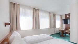 A hotel room with a bed, two windows with curtains, a desk with a chair, a flat-screen TV, and a radiator. - Regiohotel Bunte Stadt Wernigerode (Wernigerode)