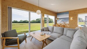A modern living room with a sofa, a chair, a wooden table, and large windows offering a view of a grassy field.