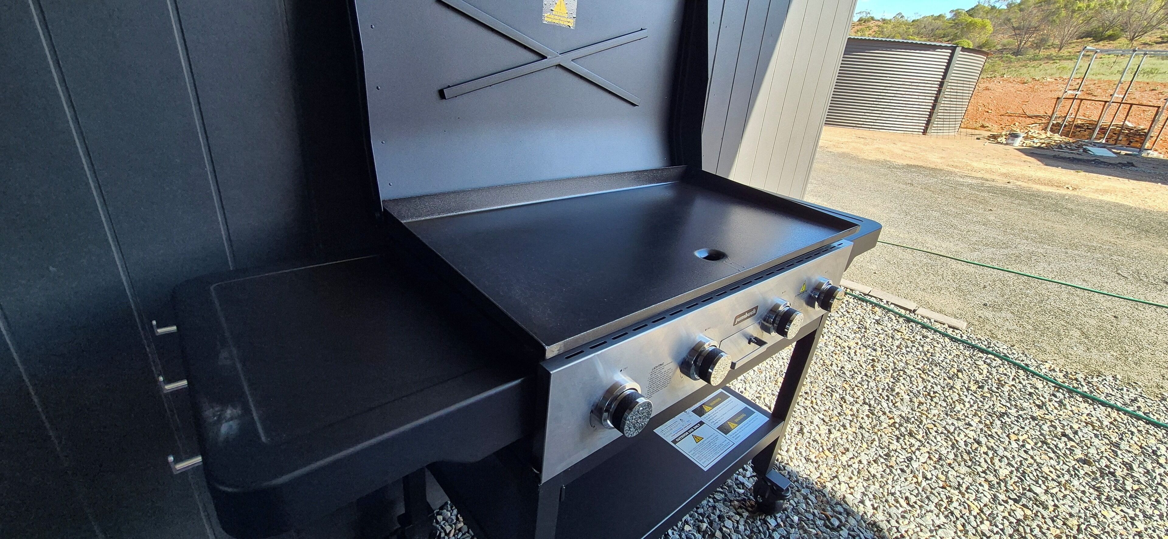 A portable grill with a black countertop and four burners, positioned outdoors on gravel.