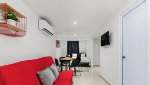 Een compacte hotelkamer met een rode bank, een kleine eettafel met stoelen, een aan de muur bevestigde televisie en een witte deur.