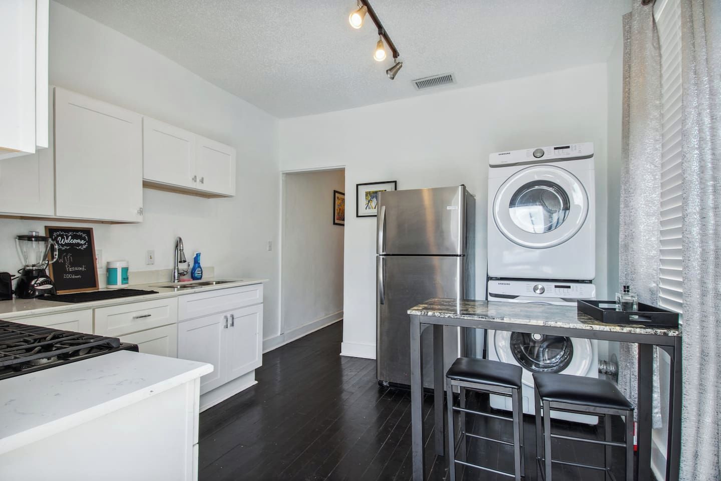 A modern kitchen with white cabinets, a stainless steel refrigerator, a washing machine, and a bar stool.