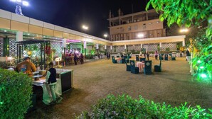 An outdoor event space with tables and chairs, a stage, and a building in the background. - Royal Mansion Lawns (Kanpur)