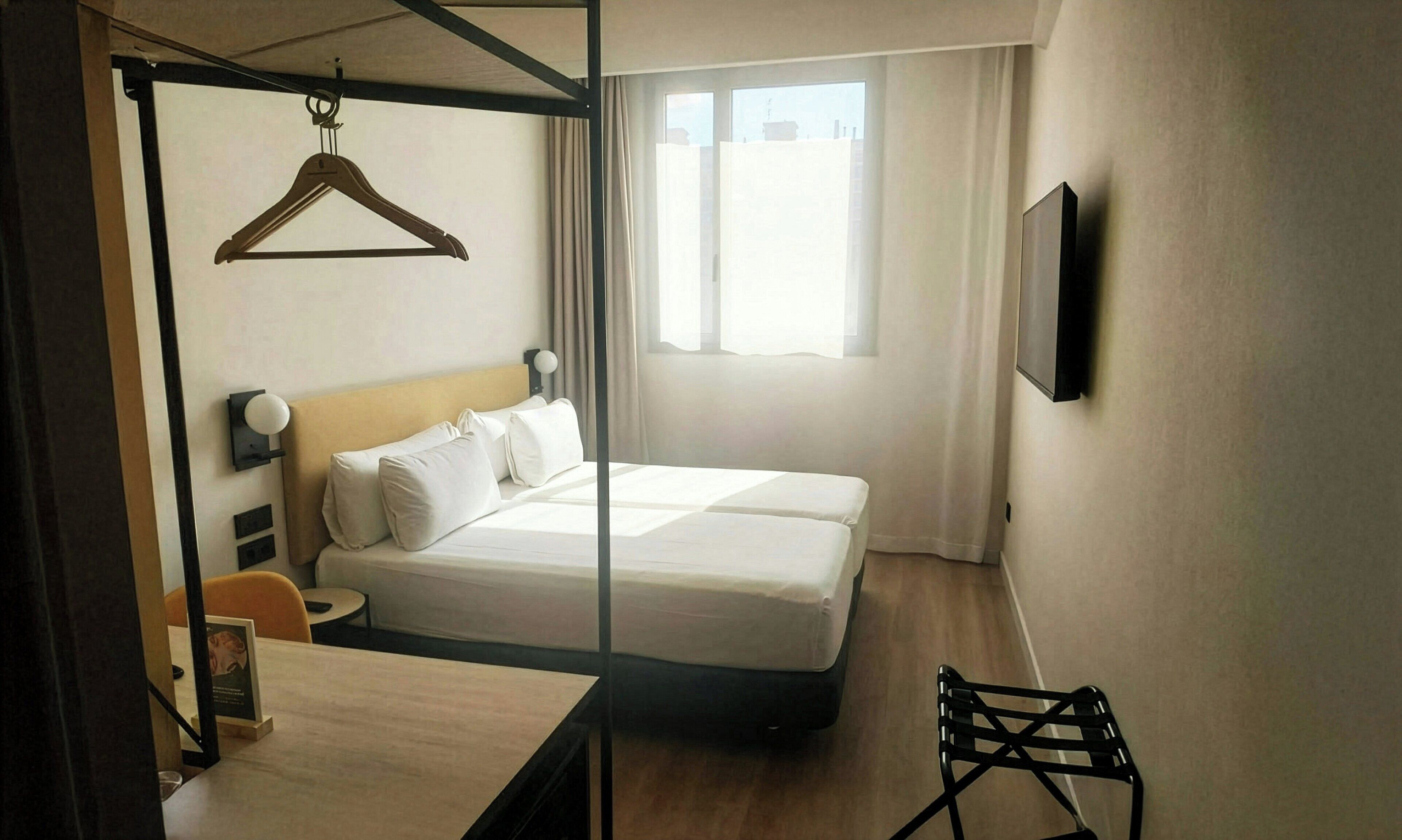 Habitación de hotel con cama, escritorio, silla, perchero y ventana con cortinas.
