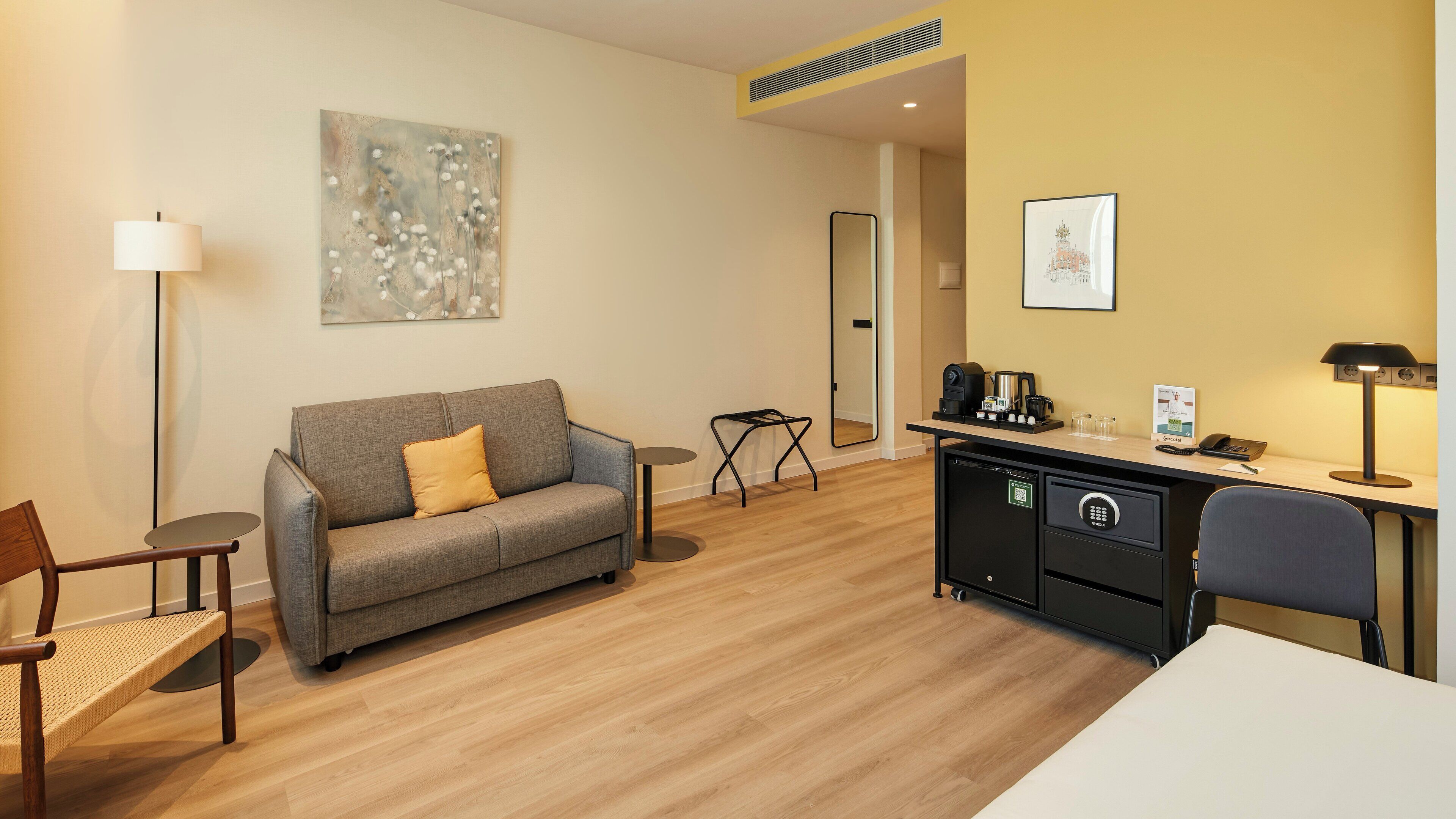A hotel room with a sofa, a small round table, a desk with a computer, a coffee machine, and a mirror.