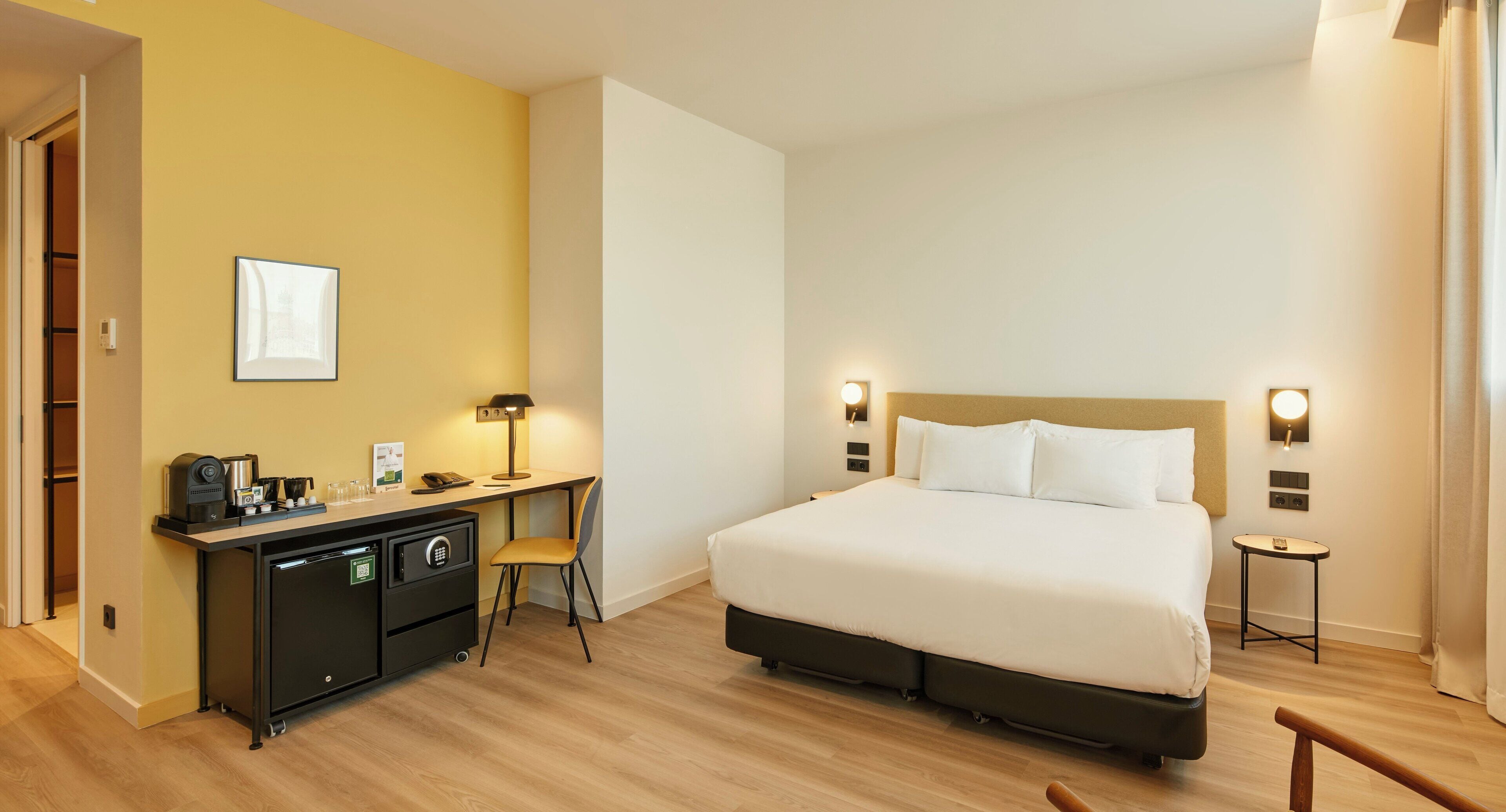 Habitación de hotel con cama, escritorio, silla y una mesita de noche con lámpara.