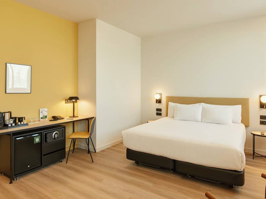 Habitación de hotel con cama, escritorio, silla y una mesita de noche con lámpara.