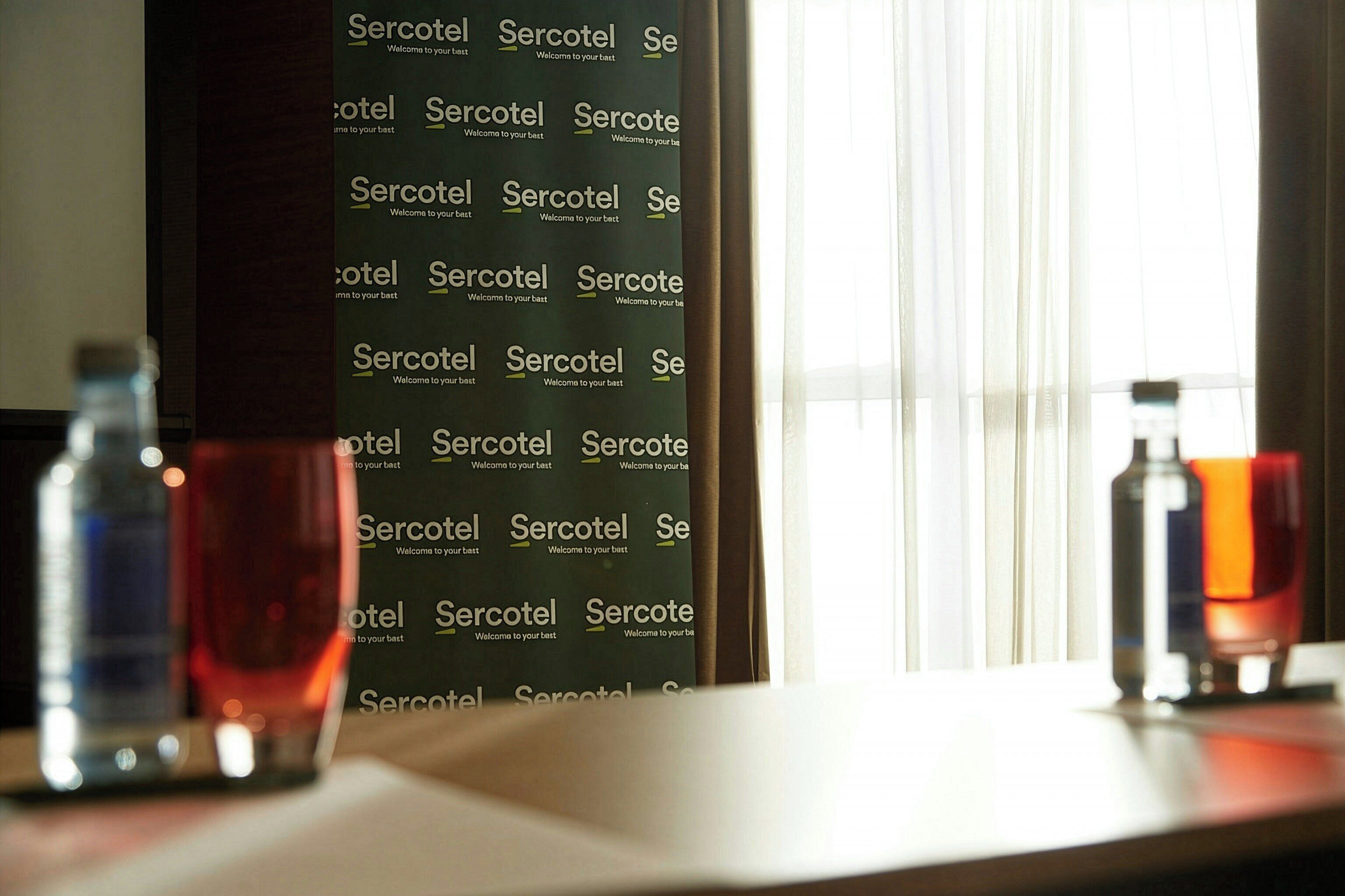 Une pièce dont un mur est orné de la mention « Sercotel » répétée, avec une fenêtre et des rideaux, et deux bouteilles posées sur une table.