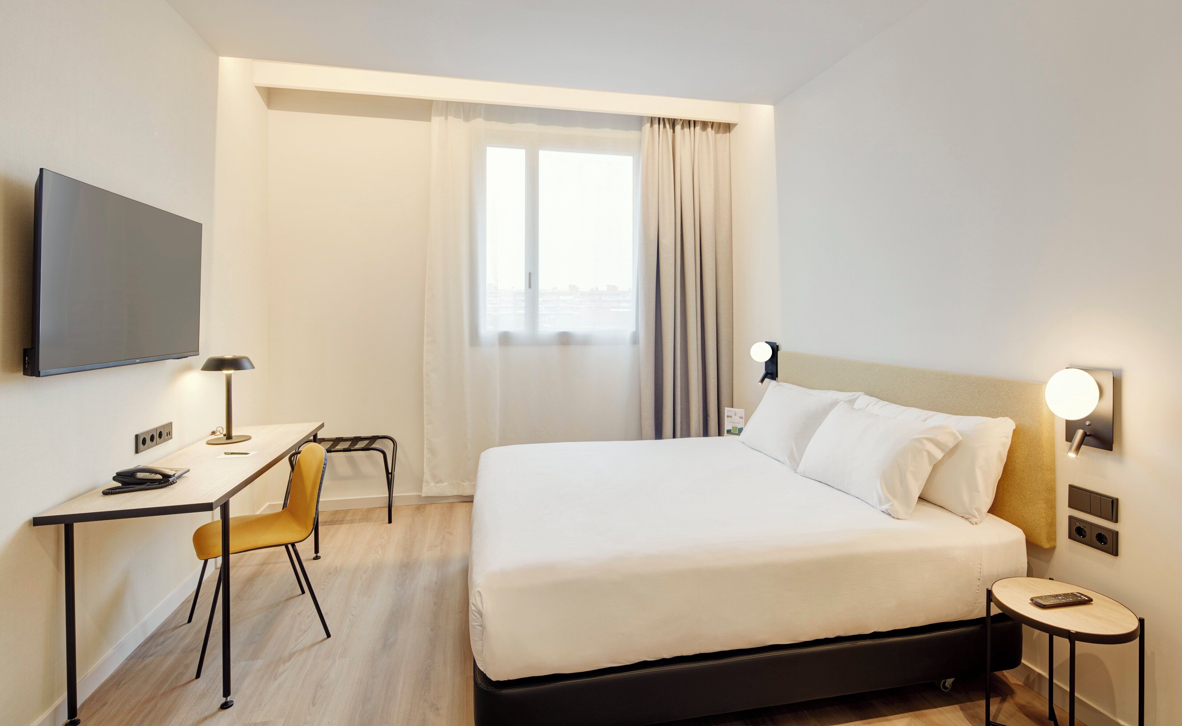 A hotel room with a bed, a desk, a chair, a TV, and a window with curtains.