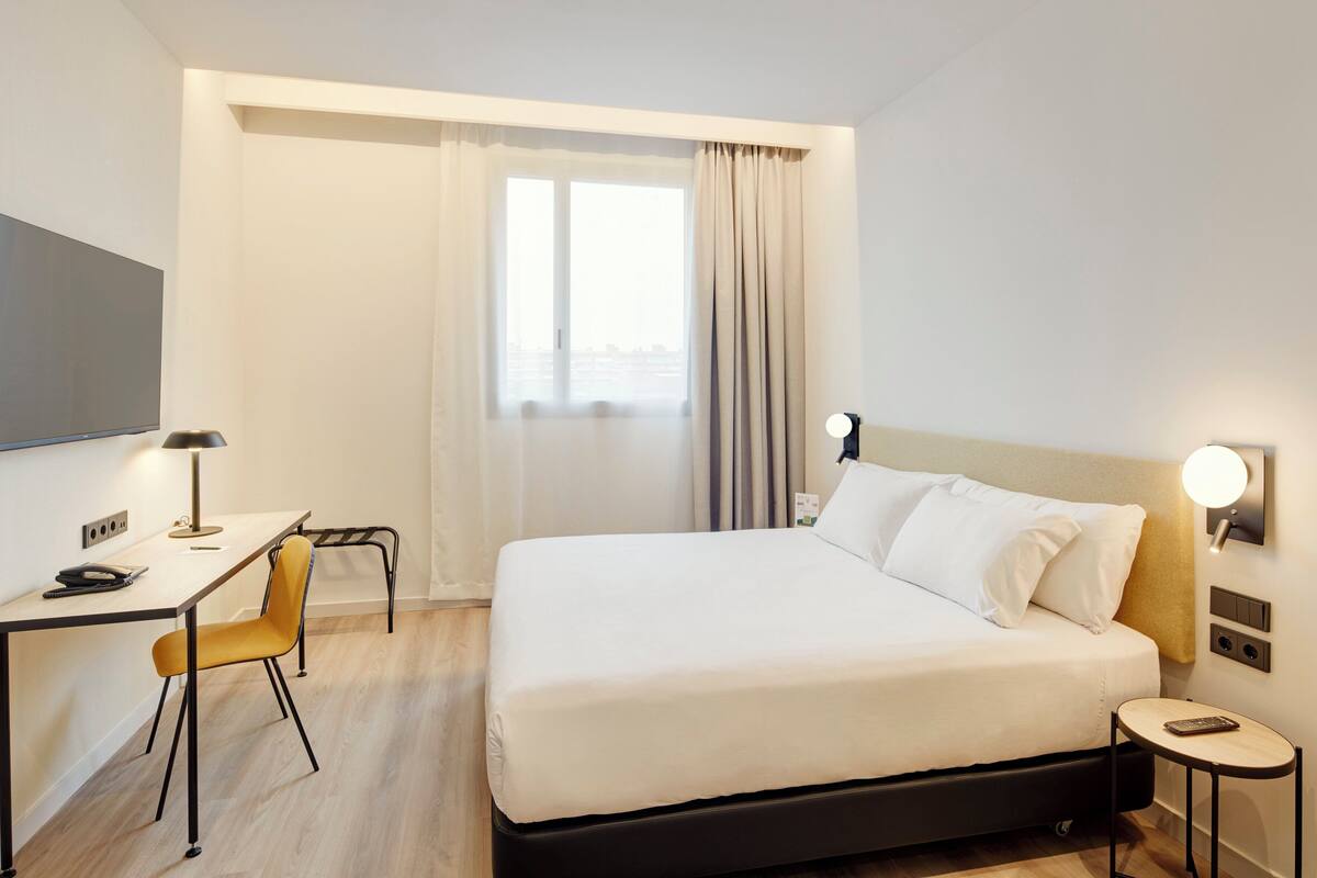 Une chambre d’hôtel avec un lit, un bureau, une chaise, une télévision et une fenêtre avec des rideaux.