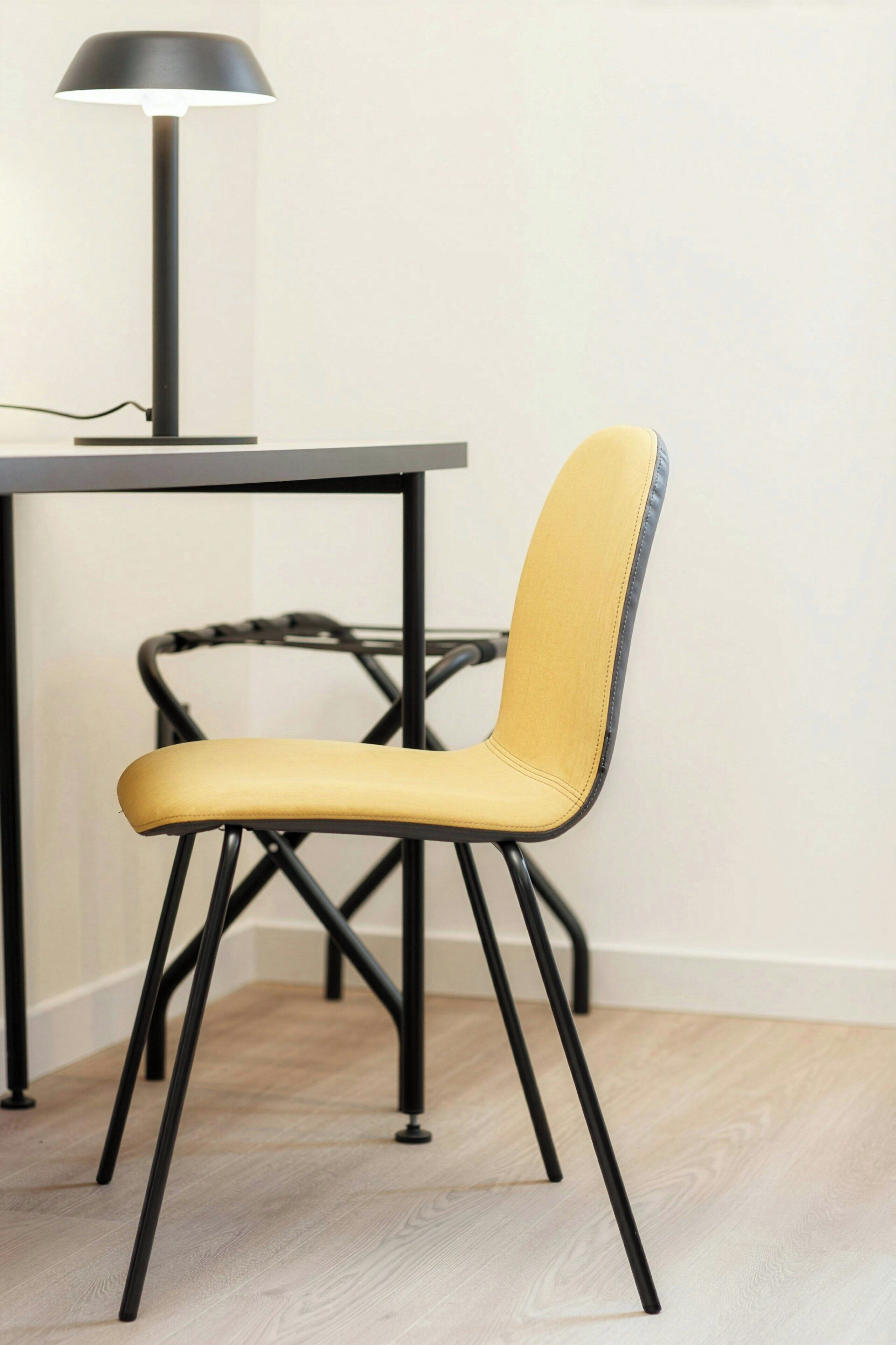 A chair with a yellow seat and black metal frame, positioned in front of a table with a black lamp.