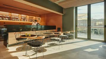 Un espace bar moderne avec un barman, des tables rondes et des chaises, ainsi qu’une grande fenêtre offrant une vue sur la ville.