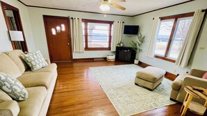 A living room with a wooden floor, beige sofa, ottoman, and a flat-screen TV mounted on the wall. - Quiet 3-bedroom house in Mexico, Kentucky (Marion)