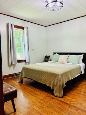 Quarto com cama, mesa de cabeceira com relógio, janela com cortinas e piso de madeira.
