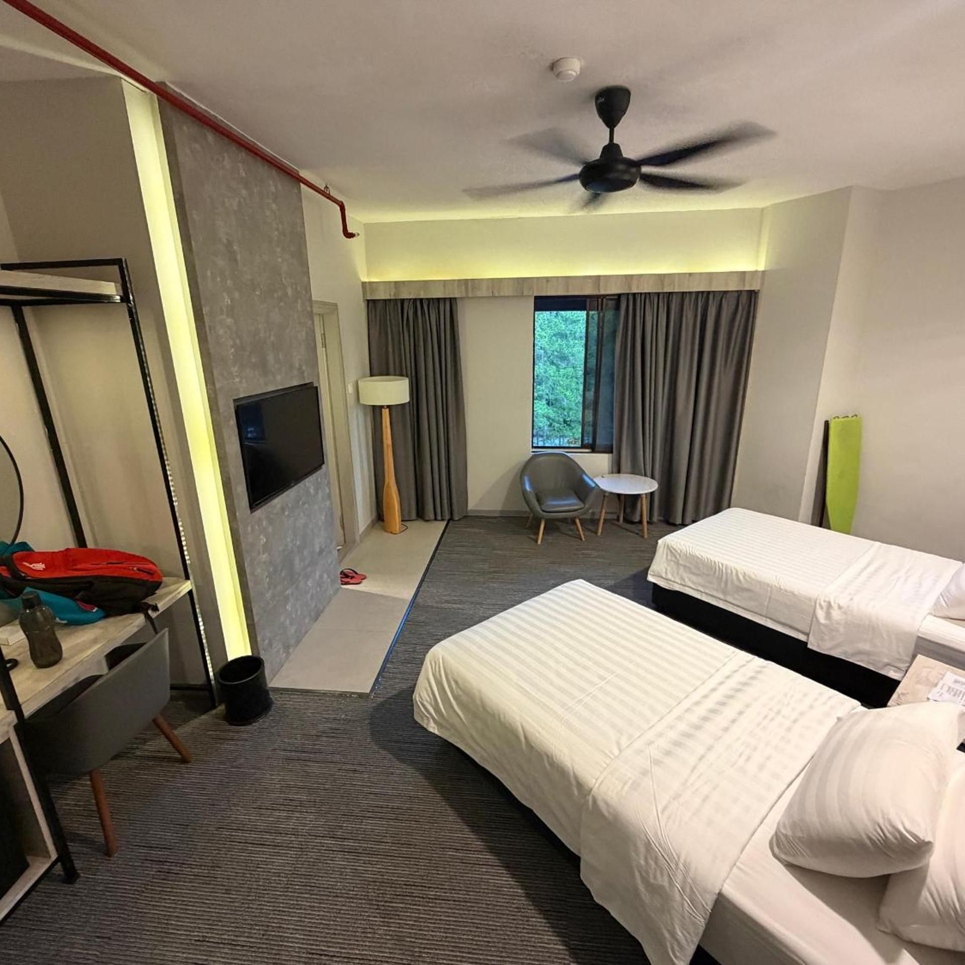 A hotel room with two beds, a desk with a TV, a chair, a small table, a ceiling fan, and a window with curtains.