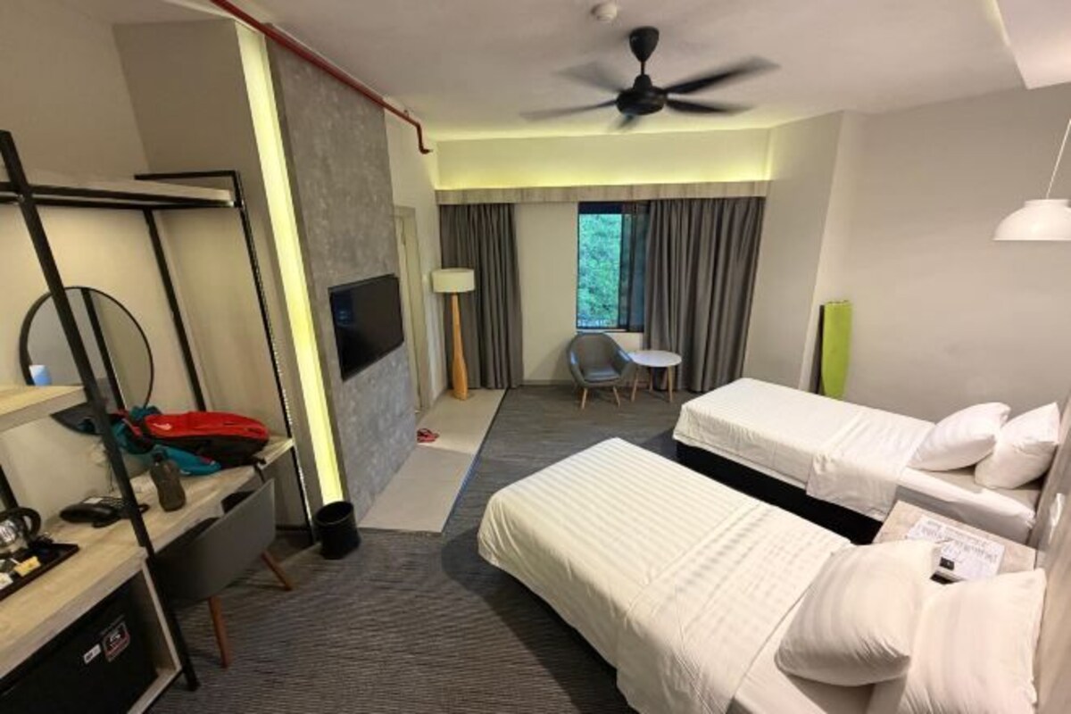 Quarto de hotel com cama, escrivaninha, cadeira, televisão e janela com cortinas.