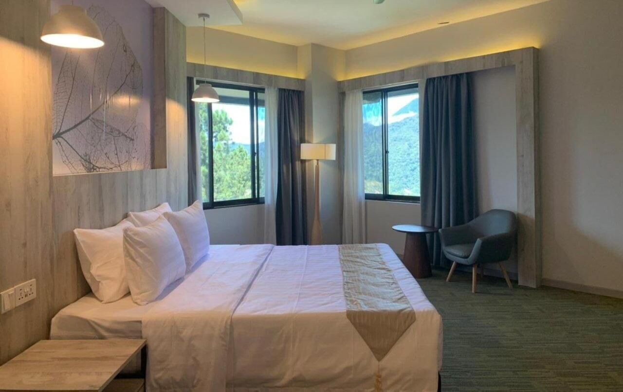A hotel room with a bed, a chair, a lamp, and large windows.