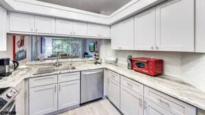 A modern kitchen with white cabinetry, a granite countertop, a red microwave, and a view of a cityscape through the window. - NAPLES 2B/2B IMPERIAL CONDO W/ STYLE & LAKE VIEWS! POOLS! NEAR EVERYTHING! 102 (Naples)