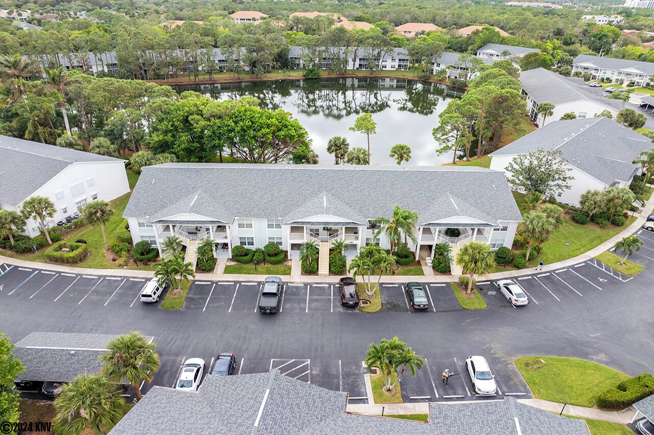 Luchtbeeld van een meerhoog hotel met een parkeerplaats ervoor, omringd door groen en een waterpartij.