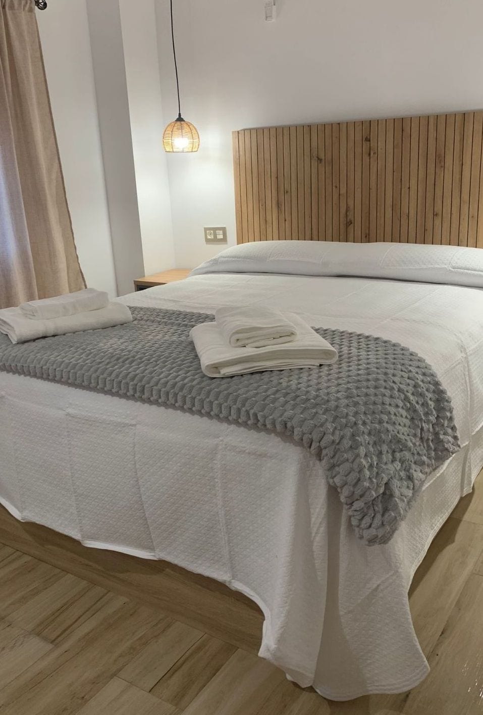 A neatly made bed with a grey blanket and white towels, a wooden headboard, and a hanging light fixture.