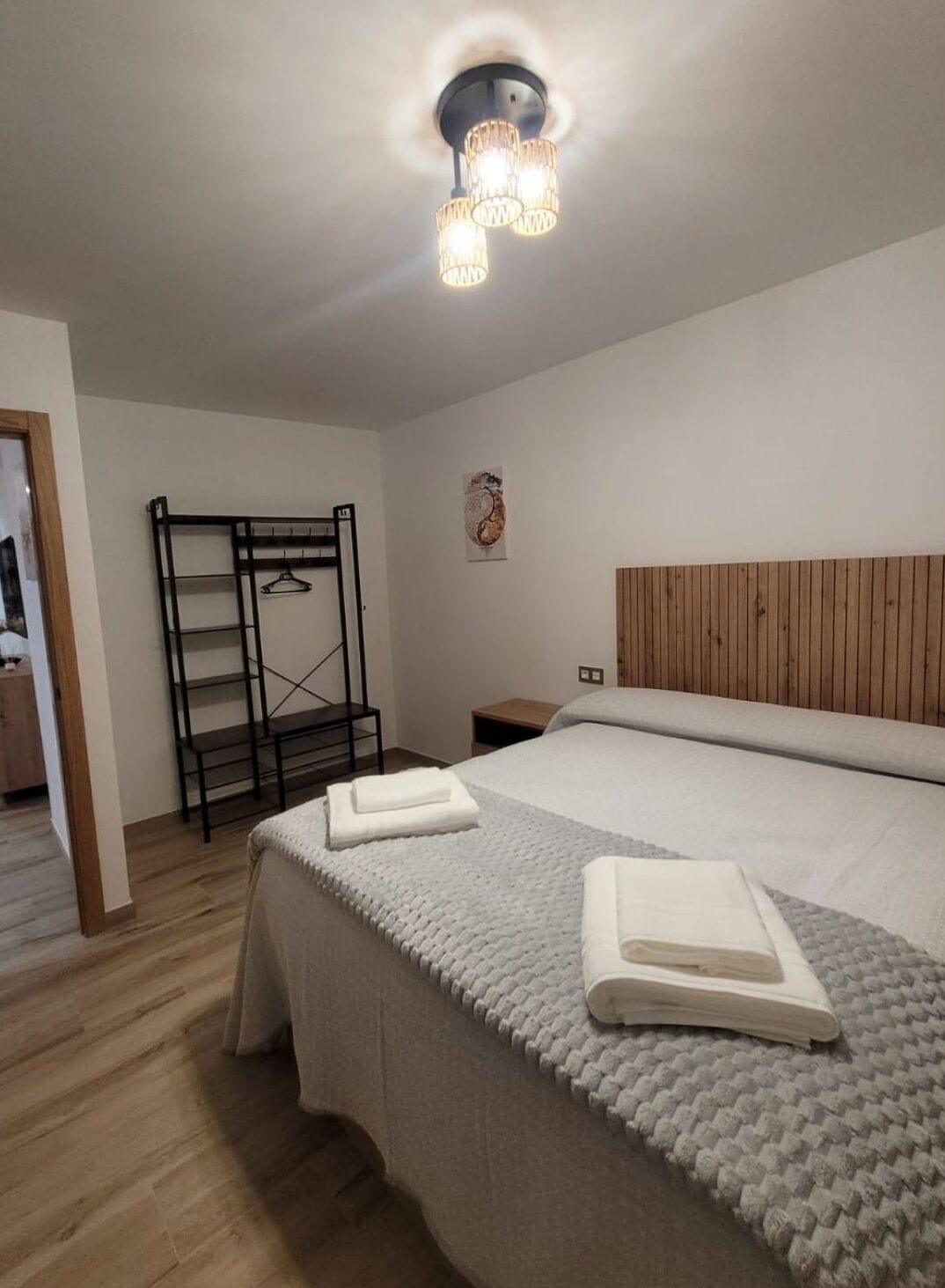 Una habitación de hotel moderna con una cama grande, cabecera de madera, una mesita de noche pequeña, un estante montado en la pared y una lámpara del techo.
