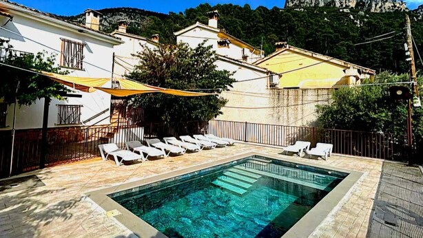 Une piscine entourée de chaises longues, bordée de bâtiments et avec une chaîne de montagnes en arrière-plan.
