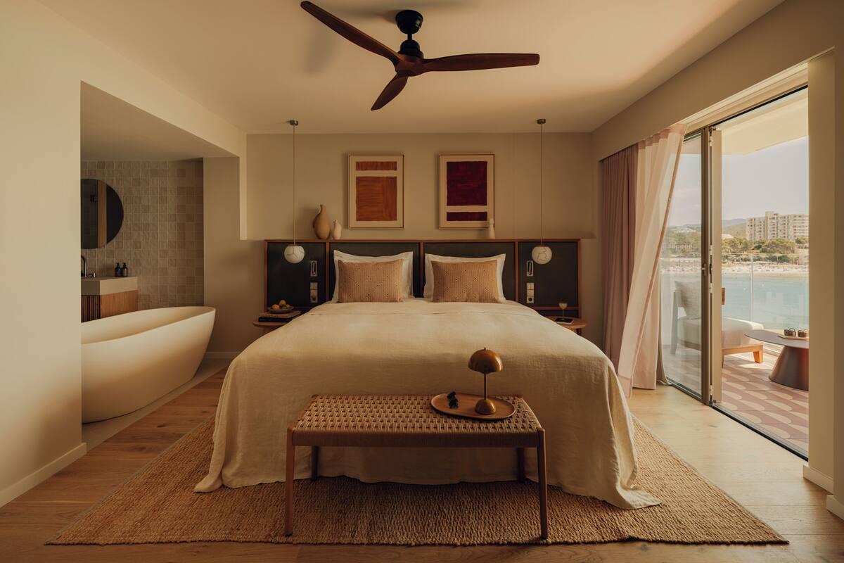 A modern bedroom with a large bed, a ceiling fan, a view of the ocean, and a bathroom with a bathtub.