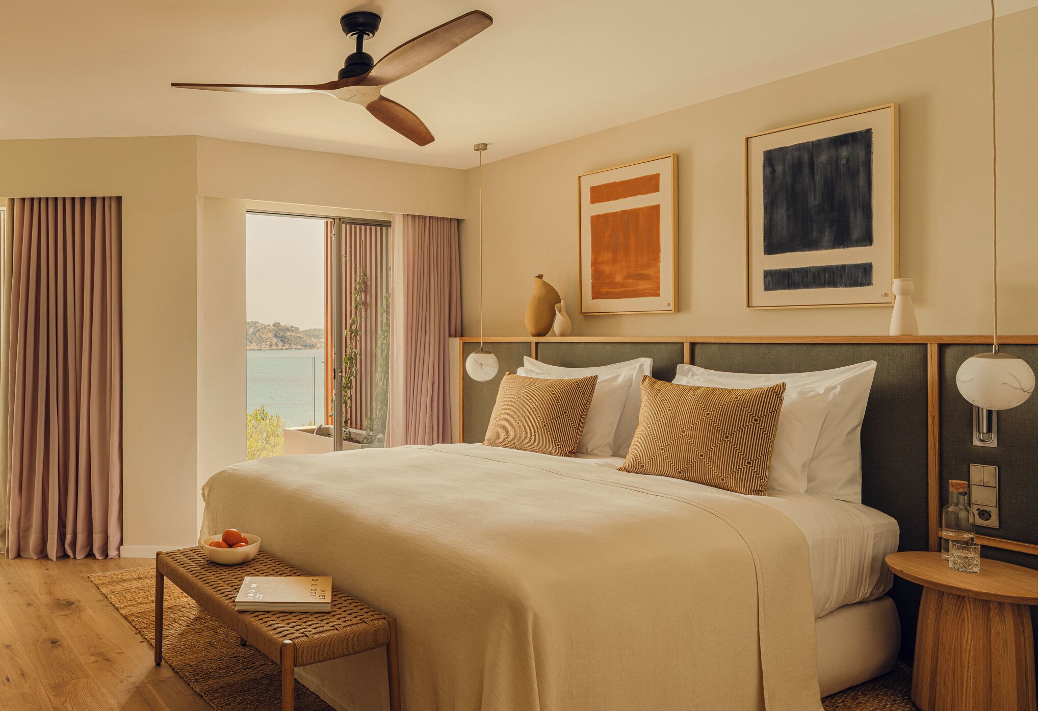 A hotel room with a large bed, a ceiling fan, a view of the sea, a bedside table with a lamp, and a framed artwork on the wall.