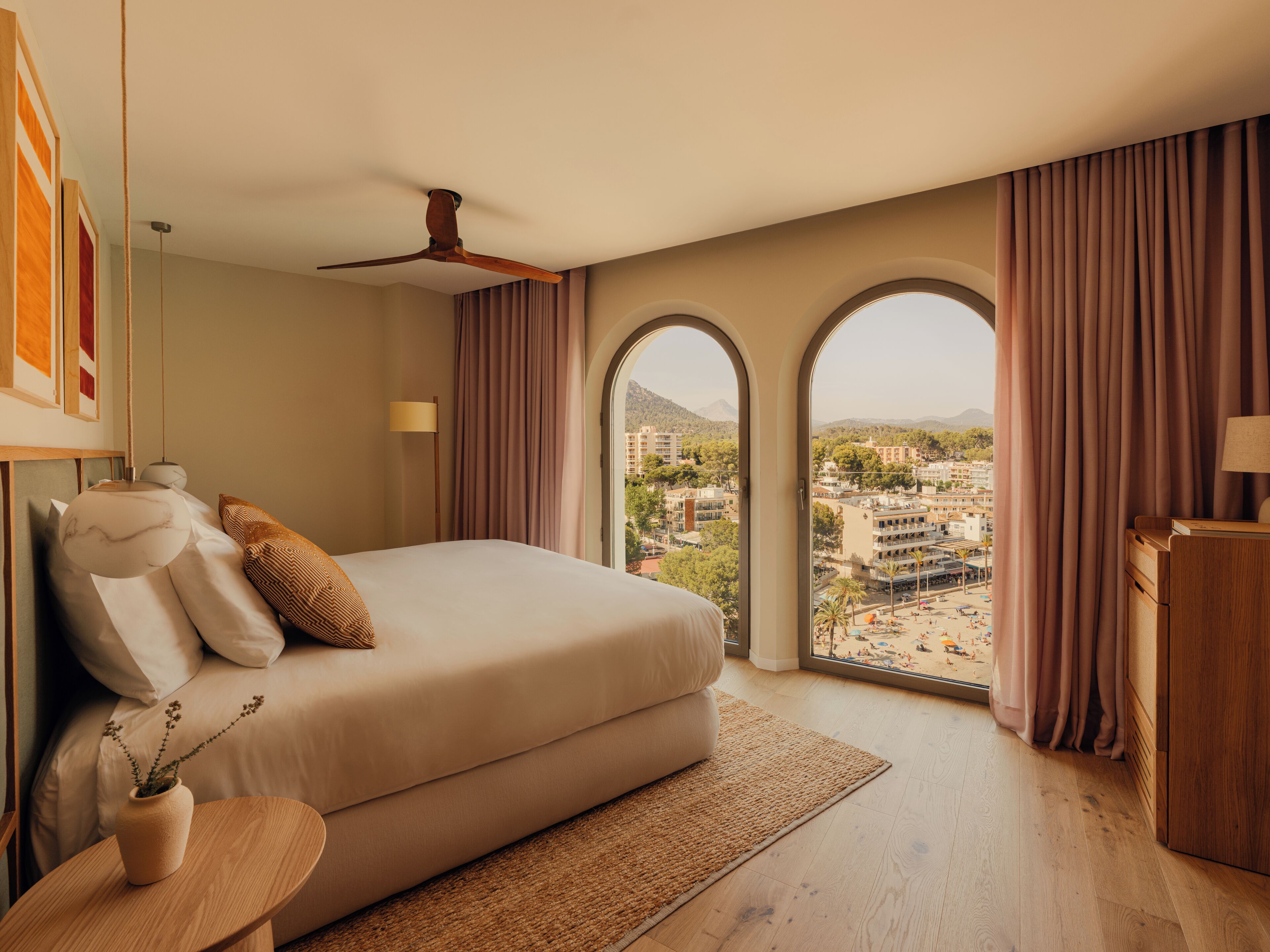 Ein Schlafzimmer mit einem großen Bett, einem Deckenventilator, Blick auf eine Stadtkulisse durch Bogenfenster und einem Nachttisch aus Holz mit einer Lampe.