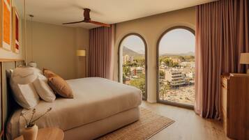 A bedroom with a large bed, a ceiling fan, a view of a cityscape through arched windows, and a wooden nightstand with a lamp.